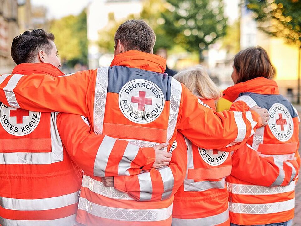 Vier ehrenamtliche Helferinnen und Helfer umarmen sich und laufen gemeinsam in Rotkreuzjacken auf einer Straße. Die Gruppe ist von hinten sichtbar.
